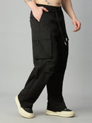 A person wearing loose black cargo pants with a drawstring waist, shown from the waist down against a light gray studio background. The pants have large flap cargo pockets and side pockets, paired with cream platform sneakers.