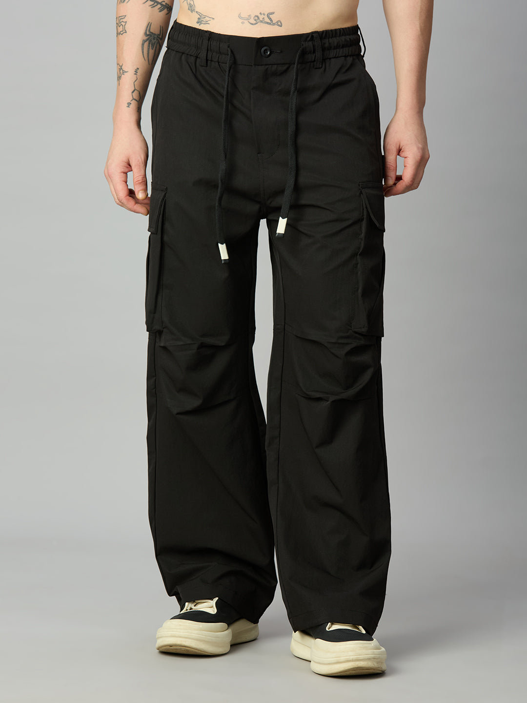 A person wearing black wide-leg cargo pants with a drawstring waist and large side cargo pockets, paired with cream sneakers, photographed against a plain light gray studio background.