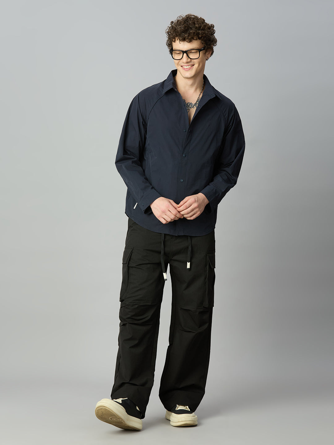 A man wearing a dark navy long-sleeve button-up shirt and black cargo pants, standing against a solid gray studio background.