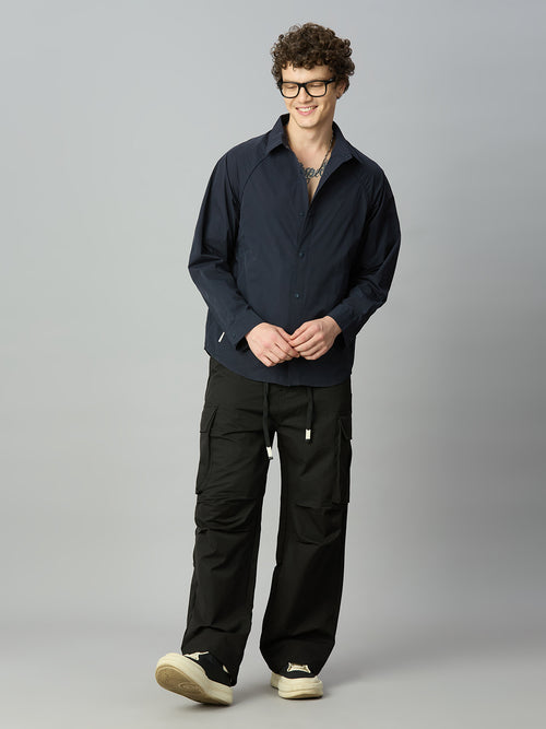 A man wearing a dark navy long-sleeve button-up shirt and black cargo pants, standing against a solid gray studio background.